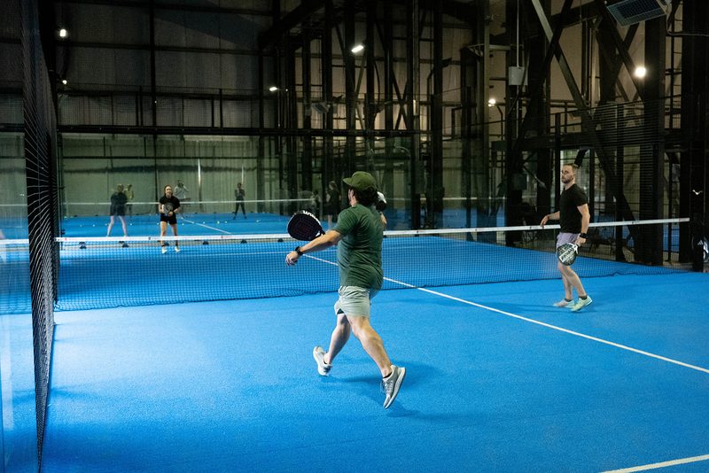 A rally seen the length of the court from behind the back glass at Foundry Padel