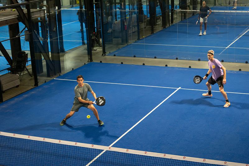 Two pairs rally across the net on a glass-walled court
