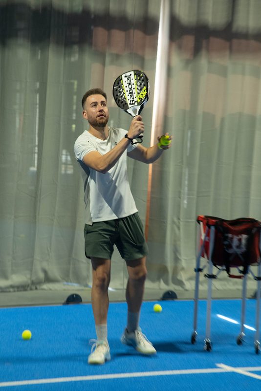 A player lines up an overhead during a coaching drill, ball basket courtside at Foundry Padel