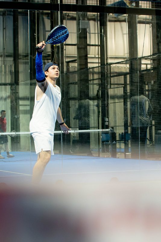 A player takes a high volley in front of the glass back wall at Foundry Padel