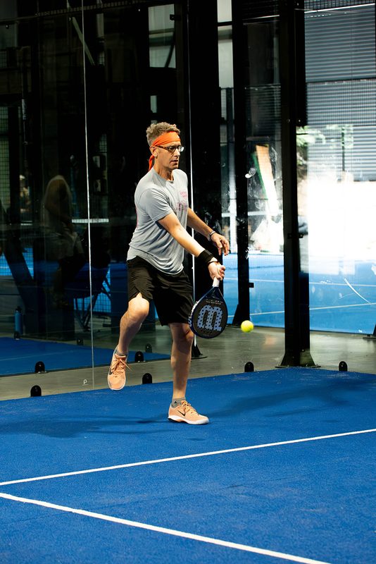 A player reaches low for the ball beside the open rollup door at Foundry Padel