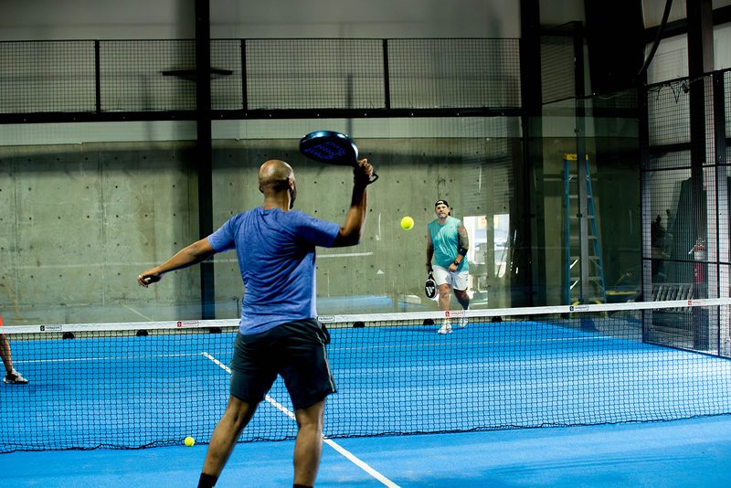 A player sets up an overhead as their opponents wait beyond the net at Foundry Padel