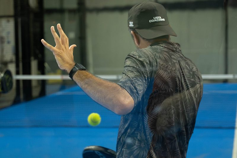 A player releases the ball at the top of the toss, seen from behind, at Foundry Padel
