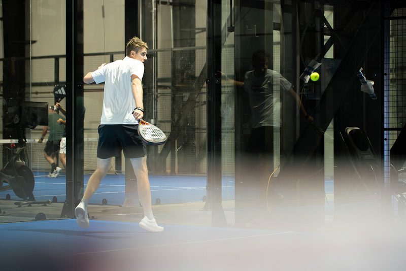 A player mid-swing, seen through the glass wall of the court at Foundry Padel